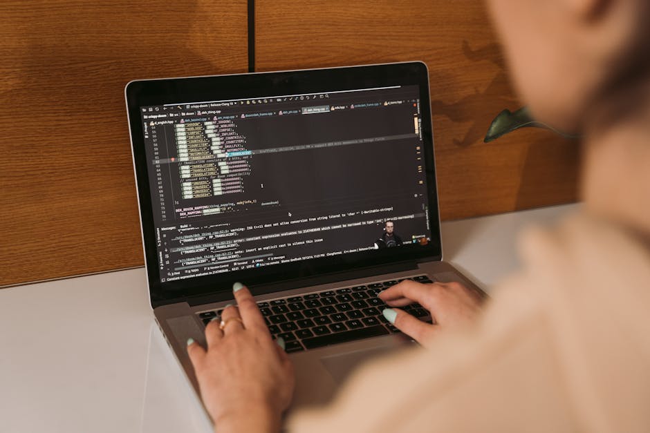 A person working on a laptop positioned on a white desk against a wooden background, with the screen displaying lines of programming code. The individual’s hands are on the keyboard, with nails pain
