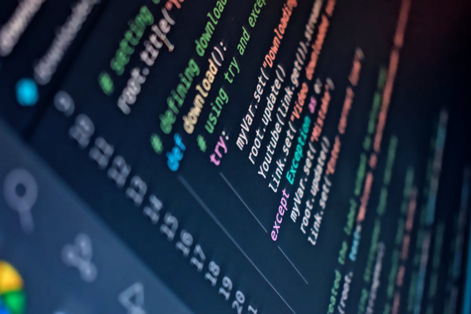 A person working on a laptop positioned on a white desk against a wooden background, with the screen displaying lines of programming code. The individual’s hands are on the keyboard, with nails pain