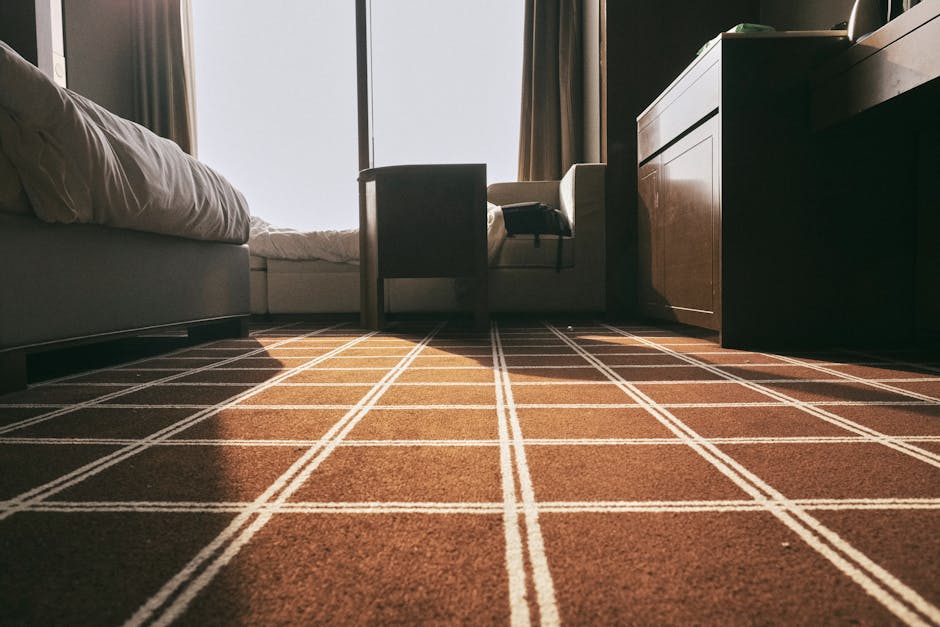 A yellow handheld vacuum cleaner with a flexible hose is placed on a traditional patterned area rug on a wooden floor in a living room. The vacuum cleaner's hose is extended and resting on the rug, in